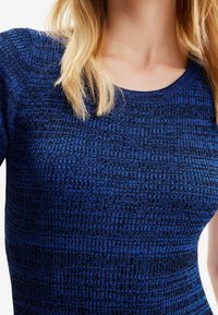 Haut tricoté bleu texturé avec un col rond. Le tissu présente un motif géométrique, créant une profondeur visuelle avec différentes nuances de bleu.