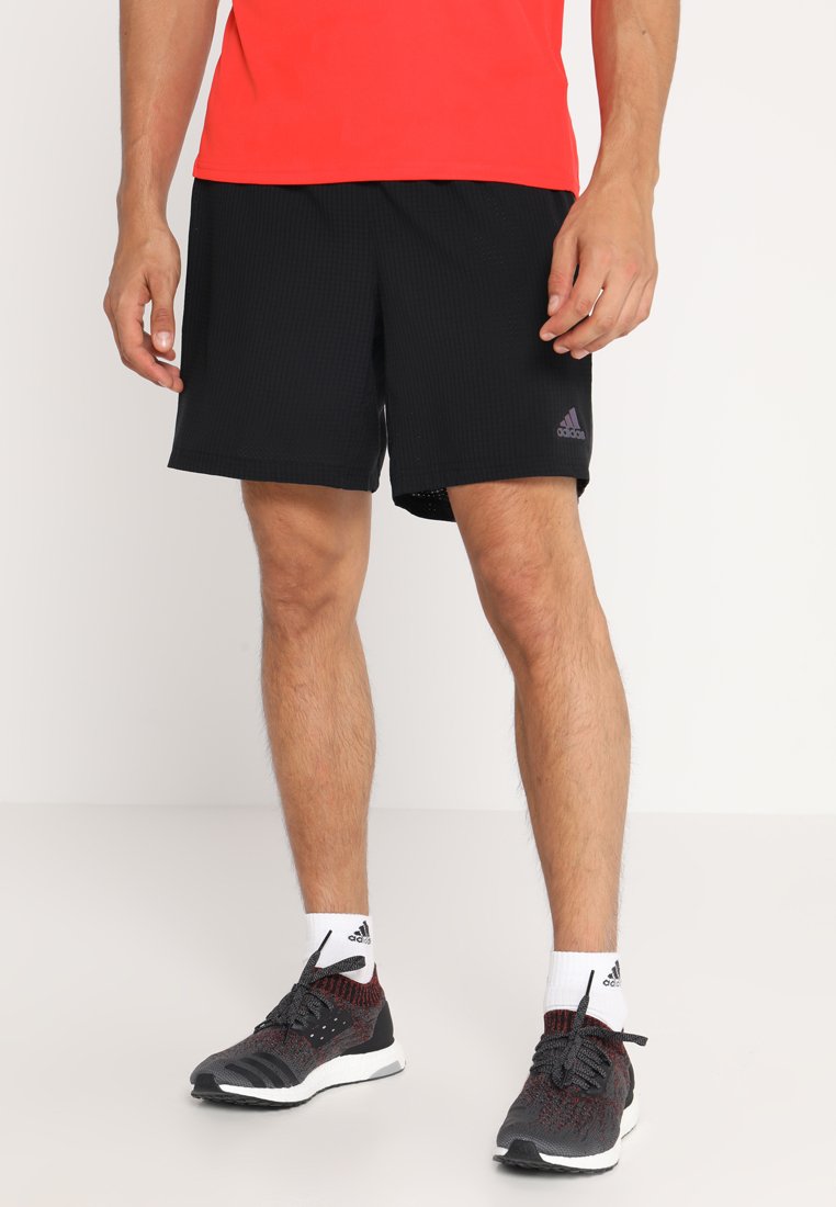 Pantalones cortos deportivos negros con una tela texturizada, que cuentan con una cinturilla elástica y un pequeño logo de Adidas en púrpura. Combinados con zapatillas negras y rojas.