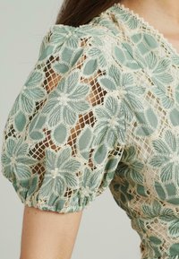 Top de encaje floral verde con mangas abullonadas, que presenta un intrincado bordado y un delicado diseño de malla. Puños elásticos que mejoran el ajuste.