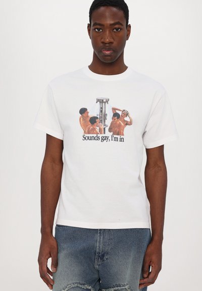 Jeune homme portant un T-shirt blanc avec une image de quatre hommes torse nu autour d'une douche et le texte "Sounds gay, I'm in."