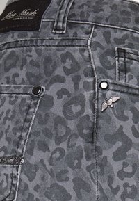 Denim gris avec motif léopard foncé, poche avec rivets métalliques, un petit emblème ailé et une étiquette noire avec texte blanc sur la ceinture.