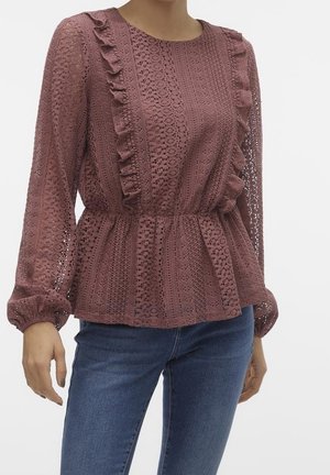 Vrouw die een mauve kanten blouse met lange mouwen en ruches en een blauwe spijkerbroek draagt, getoond van hals tot heupen op een witte achtergrond.
