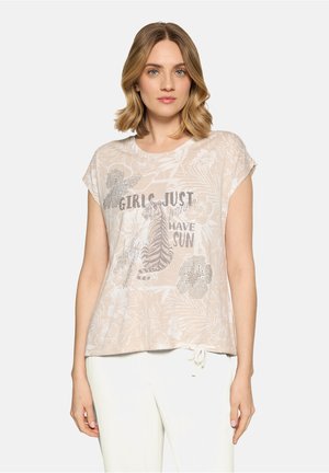 Femme portant un t-shirt beige à imprimé floral avec un tigre et le texte "Les filles veulent juste avoir du soleil", associée à un pantalon blanc, debout sur fond blanc.