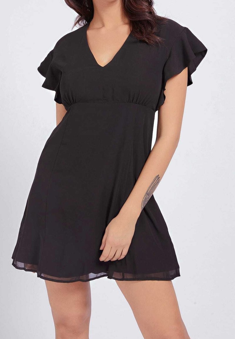 Femme portant une courte robe noire à col en V avec des manches volantées, debout, une main touchant l'ourlet de la robe.