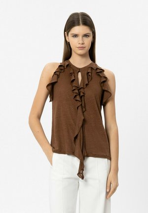 Junge Frau trägt ein ärmelloses braunes Oberteil mit Rüschen und weiße Hose, steht mit einer Hand in der Tasche vor einfarbigem Hintergrund.