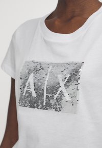 Biała bawełniana koszulka z krótkim rękawem, z okrągłym dekoltem, z prostokątną aplikacją z cekinów, na której widnieją litery "A/X" w kolorze srebrnym i czarnym.