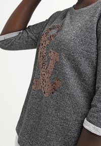Maglione grigio con maniche a tre quarti, caratterizzato da un design logo marrone testurizzato al centro e polsini bianchi a contrasto.