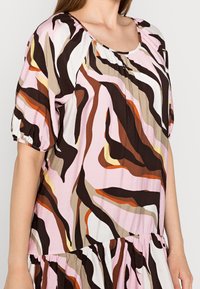 Mujer vestida con un vestido corto de mangas cortas y holgado, con un patrón abstracto en forma de oleadas en colores rosa, marrón, negro, blanco, naranja y beige.