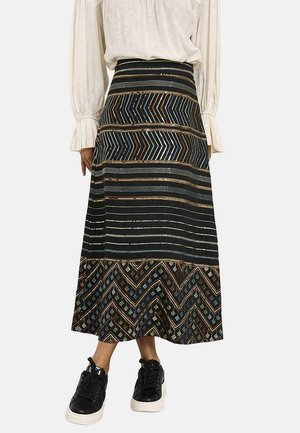 Mujer con una falda larga estampada con diseños en zigzag y rayas en negro, azul y marrón, combinada con zapatillas negras y una blusa blanca.