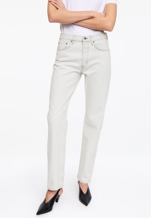 Personne portant un jean beige clair taille haute, un top blanc à manches courtes, des talons noirs pointus, bras croisés sur la poitrine, debout sur fond blanc.
