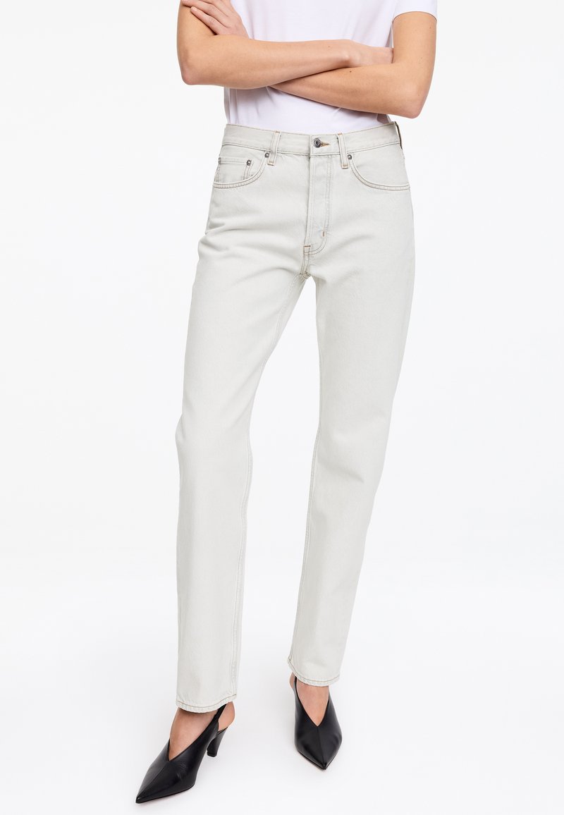 Personne portant un jean beige clair taille haute, un top blanc à manches courtes, des talons noirs pointus, bras croisés sur la poitrine, debout sur fond blanc.