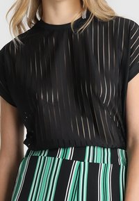 Musta läpinäkyvä toppi, jossa on pystysuorat raidat ja lyhyet hihat, yhdistettynä moniväriseen vihreä-mustaan korkeavyötäröiseen hameeseen.