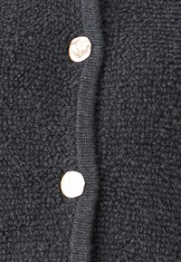 Μαύρο υφασμάτινο υλικό με απαλή, αφράτη όψη, που διαθέτει δύο στρογγυρούς χρυσούς κουμπιά κατά μήκος του μπροστινού ανοίγμ ατος για κλείσιμο.