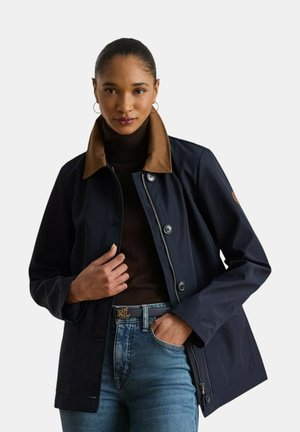 Donna con giacca blu navy con colletto marrone, dolcevita nero, jeans blu e orecchini a cerchio, che posa con una mano in tasca.