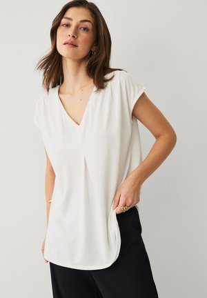 Kvinde med skulderlængde brunt hår iført en hvid kortærmet top med V-hals og sorte bukser, stående med den ene hånd i lommen.