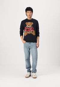 Donkerblauwe trui met een gebreid teddybeerontwerp in bruin en rood, gecombineerd met lichtblauwe jeans en witte sneakers met bruine zolen.