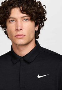 Melna polo krekls ar divu pogu aizdari, teksturētu audumu un baltu Nike logo kreisajā krūtī. Nav būtisku rakstu vai akcentu.