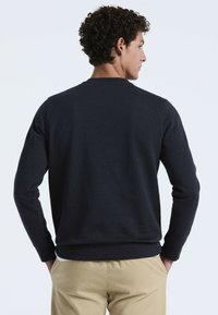 Homme avec des cheveux bouclés, portant un pull marine foncé et un pantalon beige, se tenant avec les mains dans les poches, faisant face de côté sur fond blanc.