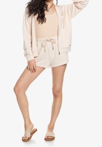 Hoodie beige corto con lacci, abbinato a un top a costine coordinato e pantaloni corti. Il modello indossa sandali a scivolo color marrone chiaro.