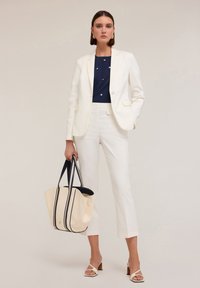 Witte blazer over een marineblauwe top met metalen accenten, gecombineerd met crème broek en een grote beige schoudertas met marineblauwe strepen. Lichte sandalen.