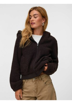 Donna con capelli lunghi biondi che indossa orecchini a cerchio d'argento, una felpa nera con zip e pantaloni beige su uno sfondo semplice.