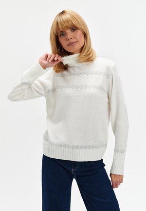 Blondynka w białym swetrze z wzorem i ciemnoniebieskich dżinsach, trzymająca kołnierz swetra jedną ręką.