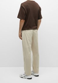 Persona che indossa pantaloni beige, sneakers bianchi e una camicia a maniche corte marrone, in piedi davanti a uno sfondo bianco uniforme.