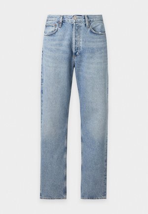 Mann trägt ein hellbeiges T-Shirt mit kurzen Ärmeln, hellblaue hoch taillierte Jeans und schwarze Schuhe, steht vor einem schlichten weißen Hintergrund.