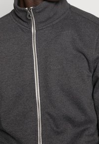 Giacca grigia con zip, dotata di un colletto alto e una zip bianca. Il tessuto è testurizzato e morbido, enfatizzando la durevolezza e il comfort.