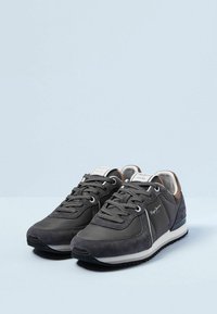 Ett par mörkgrå lediga sneakers med vita mellansole, texturerad yttersula och märkta sidor och tunga på en ljusblå yta.