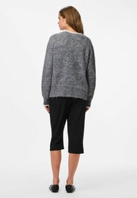 Femme de dos portant un pull en laine grise, un pantalon noir court et des chaussures noires à enfiler sur fond blanc.
