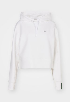 Hvit cropped hettegenser laget av bomullblanding, med snor i hetta, ribbestrikkede mansjetter, og en liten brodert logo på brystet.