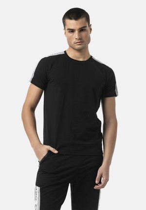 Jeune homme aux cheveux courts et foncés, portant un t-shirt noir avec des bandes blanches sur les côtés et un pantalon noir, debout avec une main dans la poche.