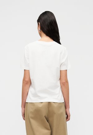 Femme aux longs cheveux foncés portant un t-shirt blanc uni à manches courtes et un pantalon ample kaki, de dos sur un fond uni.