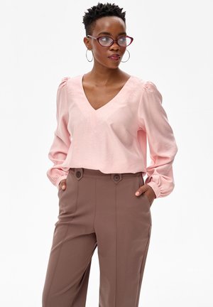 Frau mit kurzen Haaren und Brille, trägt eine rosa Bluse mit Puffärmeln und braune, hoch taillierte Hosen, Hände in den Taschen, schaut nach rechts.