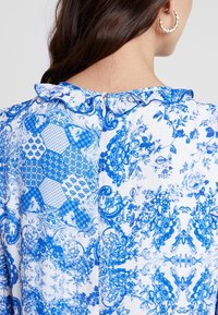 Blusa de patrón floral azul con cuello de volantes, que presenta diseños intrincados en varias tonalidades de azul sobre un fondo blanco. Cremallera en la parte posterior.