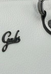 Witte leren tas met een zwart "Gabs" logo, een gladde textuur, zilverkleurige hardware en een cirkelvormig zwart detail met reliëftekst.