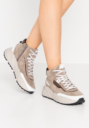 Nogi w taupe zamszowych wysokich sneakersach z białymi sznurówkami, masywnymi beżowymi podeszwami i bocznym zamkiem błyskawicznym na jasnym tle.