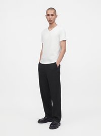 Homme debout, mains dans les poches, portant un t-shirt blanc à col en V manches courtes, pantalon noir et chaussures sombres sur fond blanc uni.