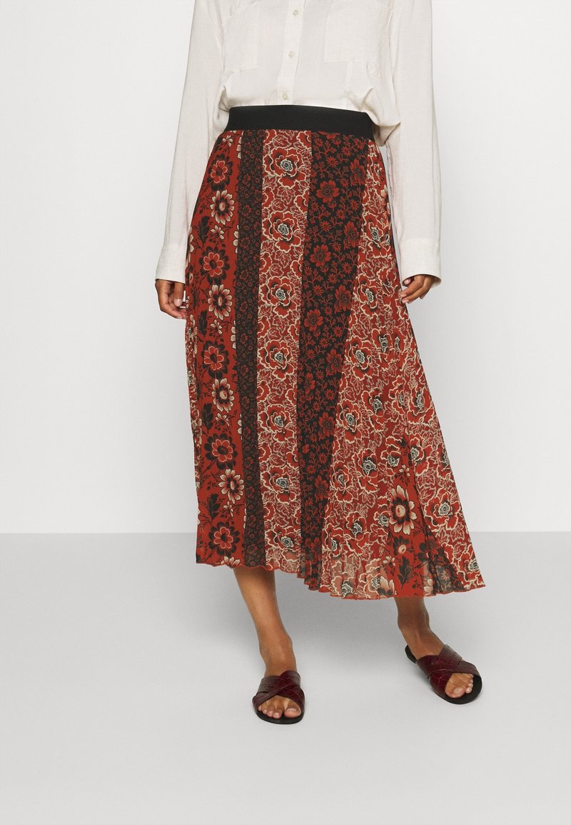 Jupe midi à motif rouge avec des designs floraux et paisley, taille élastique noire et tissu fluide. Associée à des sandales plates noires.