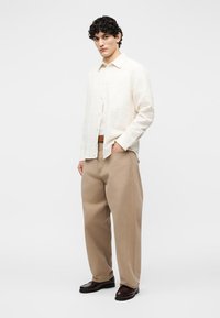 Muž s kučeravými vlasmi v krémovej košeli na gombíky, béžových širokých nohaviciach, hnedom opasku a tmavohnedých topánkach, stojaci s jednou rukou v vrecku.