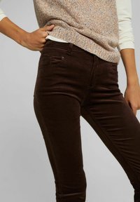Personne portant un pantalon skinny marron foncé et un pull sans manches multicolore en tricot par-dessus une chemise à manches longues blanche.