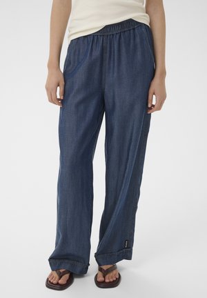Persona che indossa pantaloni di jeans blu scuro larghi con vita elastica e sandali marroni con punta quadrata, con le mani rilassate lungo i fianchi.