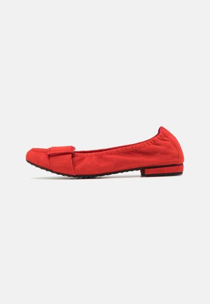 Chaussure plate en suède rouge avec un bout carré, une sangle décorative à l'avant et un petit talon, présentée en profil latéral sur un fond blanc.