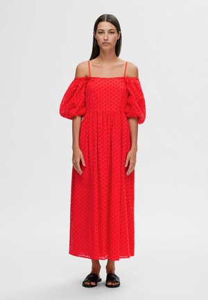 Rød off-shoulder kjole med puffærmer, struktureret stof med udskæringsmønstre og en lang, flydende nederdel, parret med sorte sandaler.