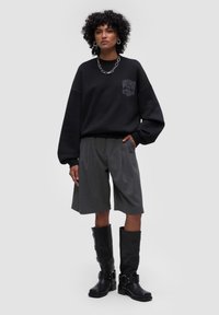 Maglione nero oversize con logo, pantaloni shorts grigi plissettati e stivali neri in pelle al ginocchio con fibbie e finitura testurizzata.