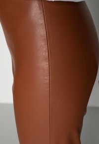 Close-up van een persoon die strakafzittende bruine leren broek draagt, met zichtbare stiknaad details langs de zijkant van het been.