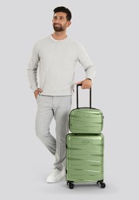 Man in een lichtgrijze outfit staat met één hand in zijn zak en de andere op de handvat van een groene koffer set, glimlachend en omhoog kijkend.