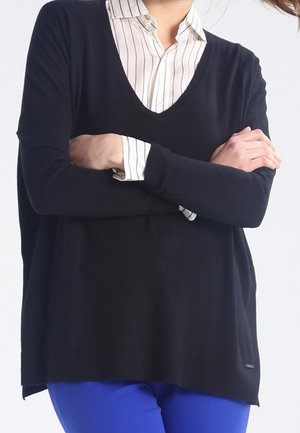 Person wearing a black V-neck sweater over a white striped shirt with arms crossed, paired with bright blue pants.
