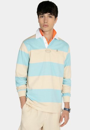 Joven con una camiseta de rugby de manga larga a rayas azules y beige con un polo de cuello naranja debajo, posando frente a un fondo blanco.